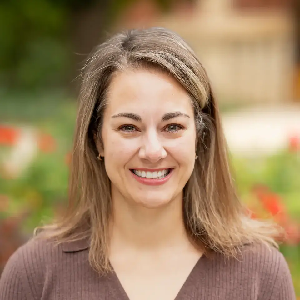 Smiling woman with shoulder-length brown hair in a brown buttoned top, standing outdoors with a blurred garden backdrop