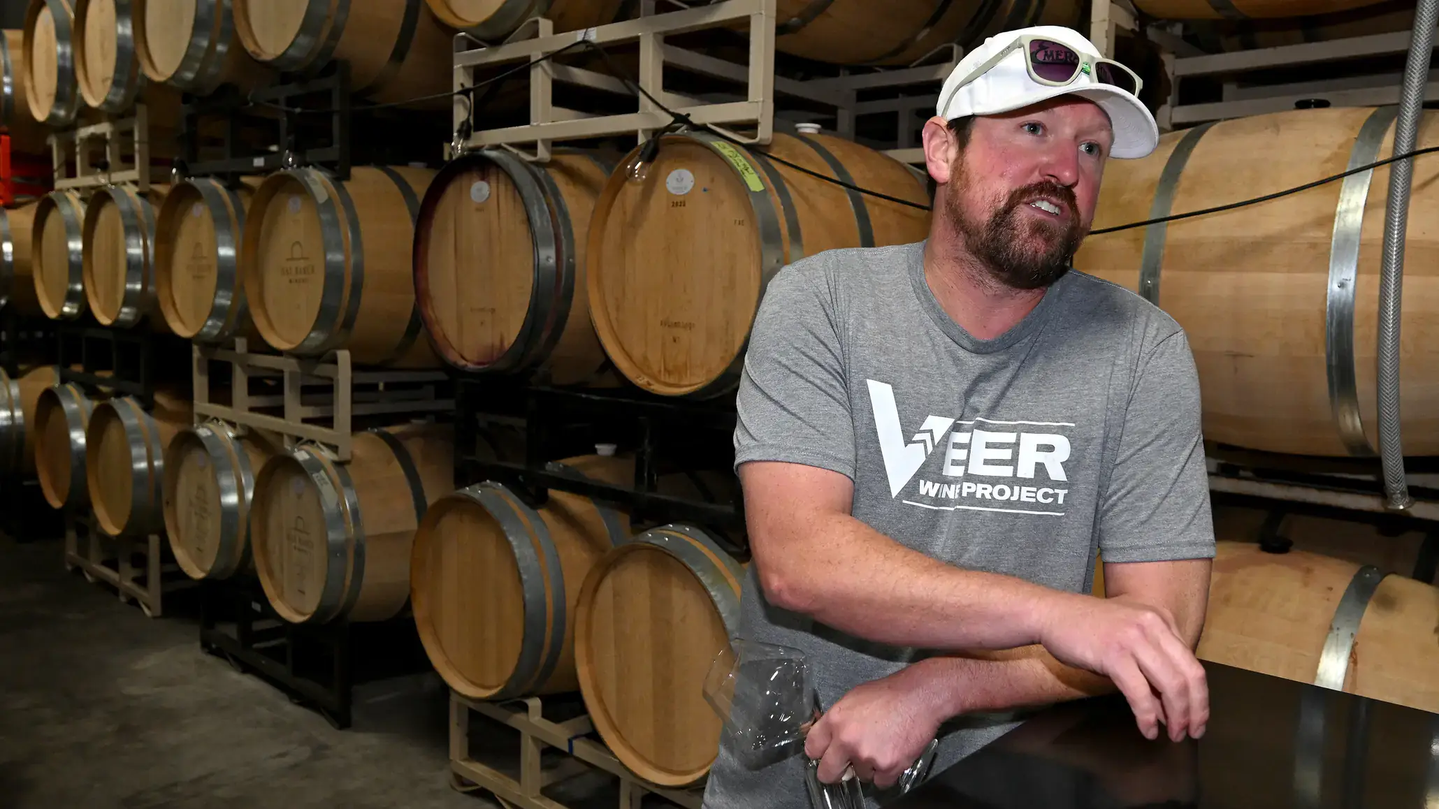 A person in a grey shirt and white cap, standing in front of stacked wine barrels in a storage area.