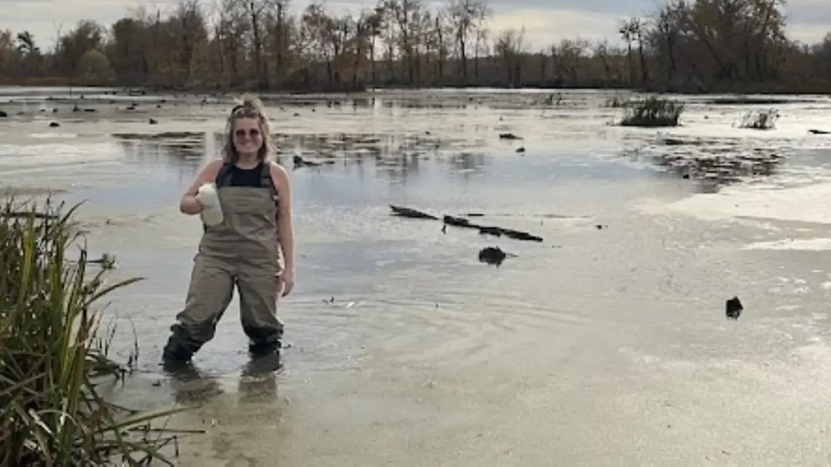 Woman wearing waders stands in a pond holding a jar of water.