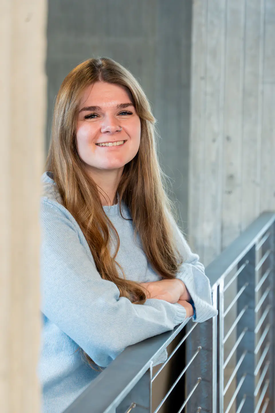 Smiling Jasmine Martinez, wearing blue sweater, leaning on a balcony rail.