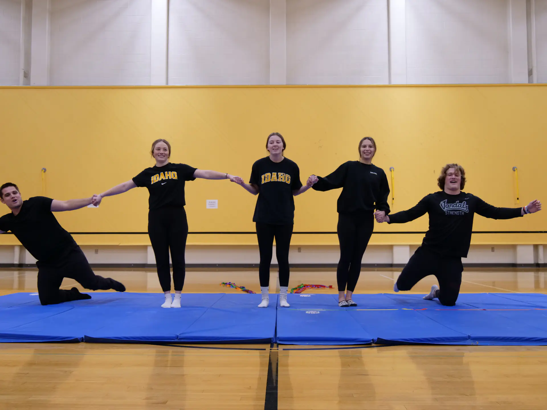 Five people on blue mats in a gymnasium, dressed in dark clothing, holding hands, performing a coordinated pose.