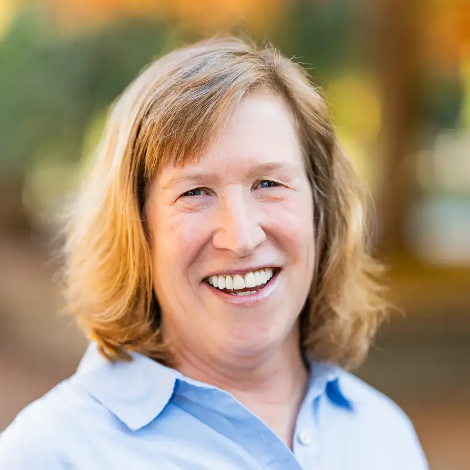 Smiling woman in a light blue button-down shirt, outdoors with a softly blurred autumn background.