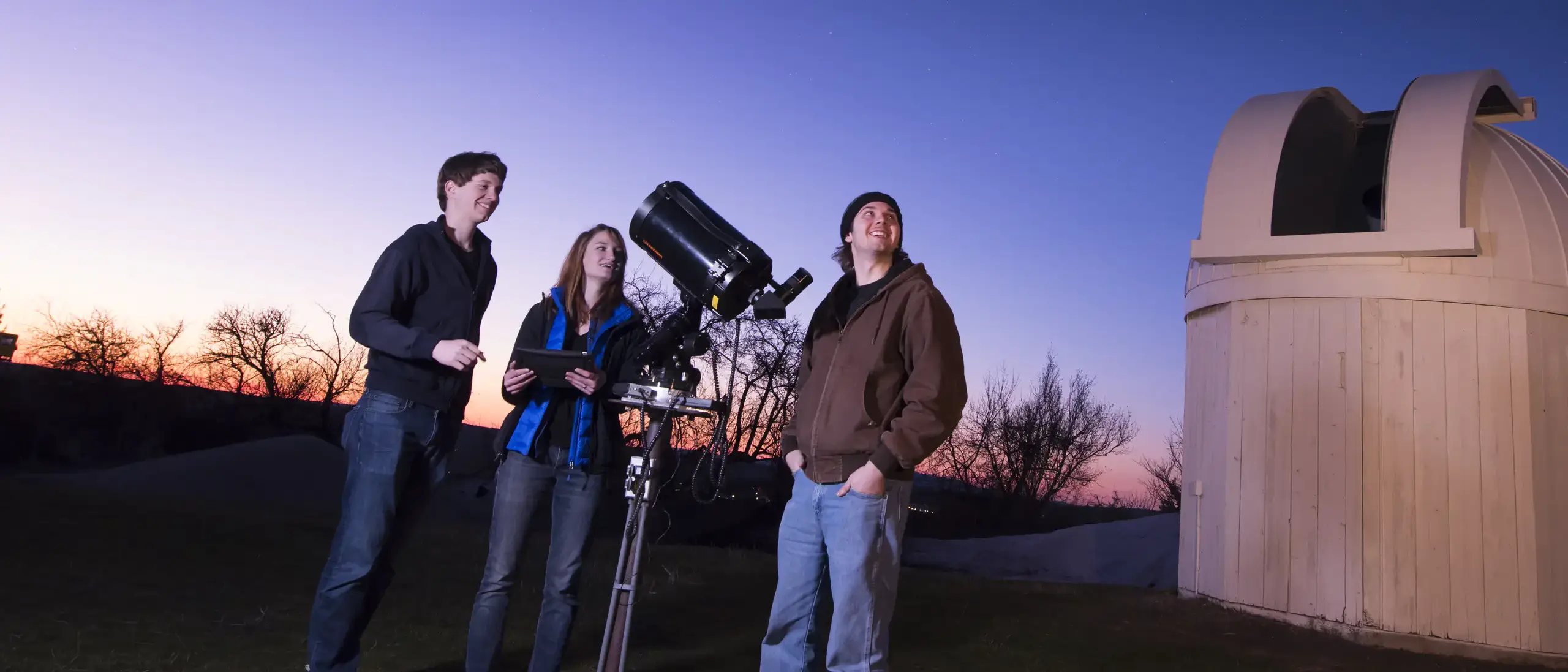 Three people stand near a telescope, observing the night sky at dusk, outside a small observatory building.