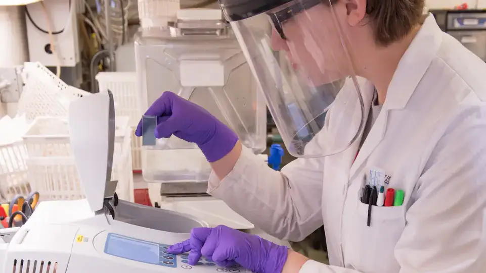A female with a protective clear face plate testing a sample in a machine.