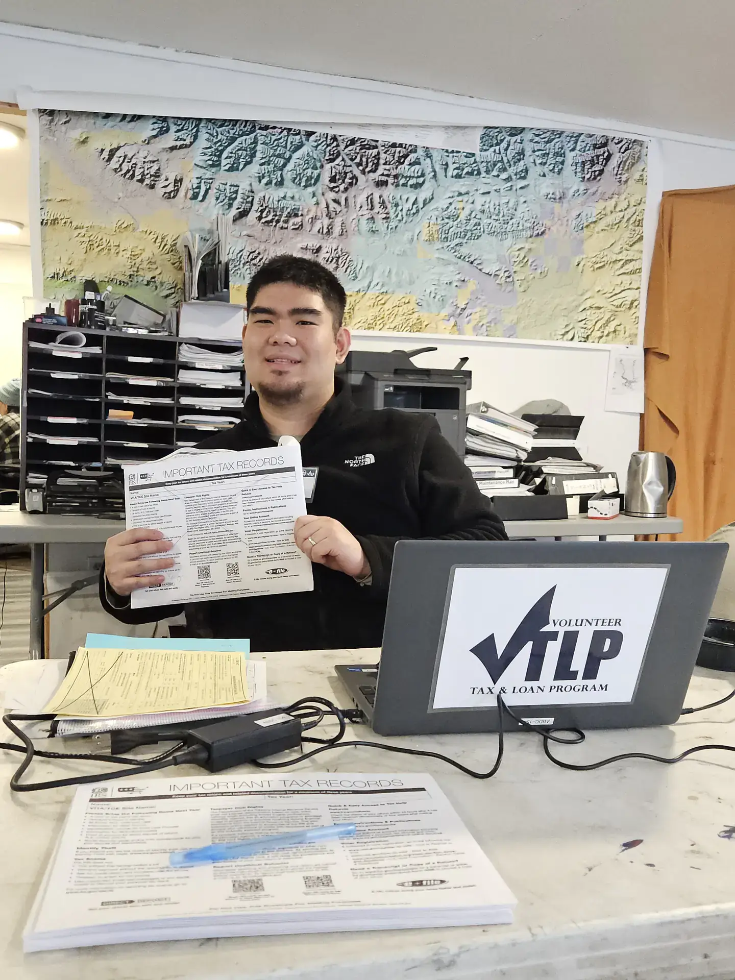 Young Sim sits at a table holding a tax preparation document. On the table are a laptop and more tax paperwork.