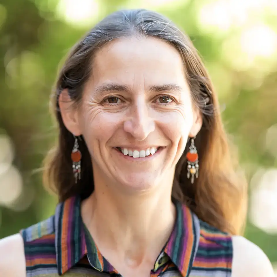 Smiling individual with long brown hair wearing a striped sleeveless shirt and earrings, outdoors with blurred greenery in the background.
