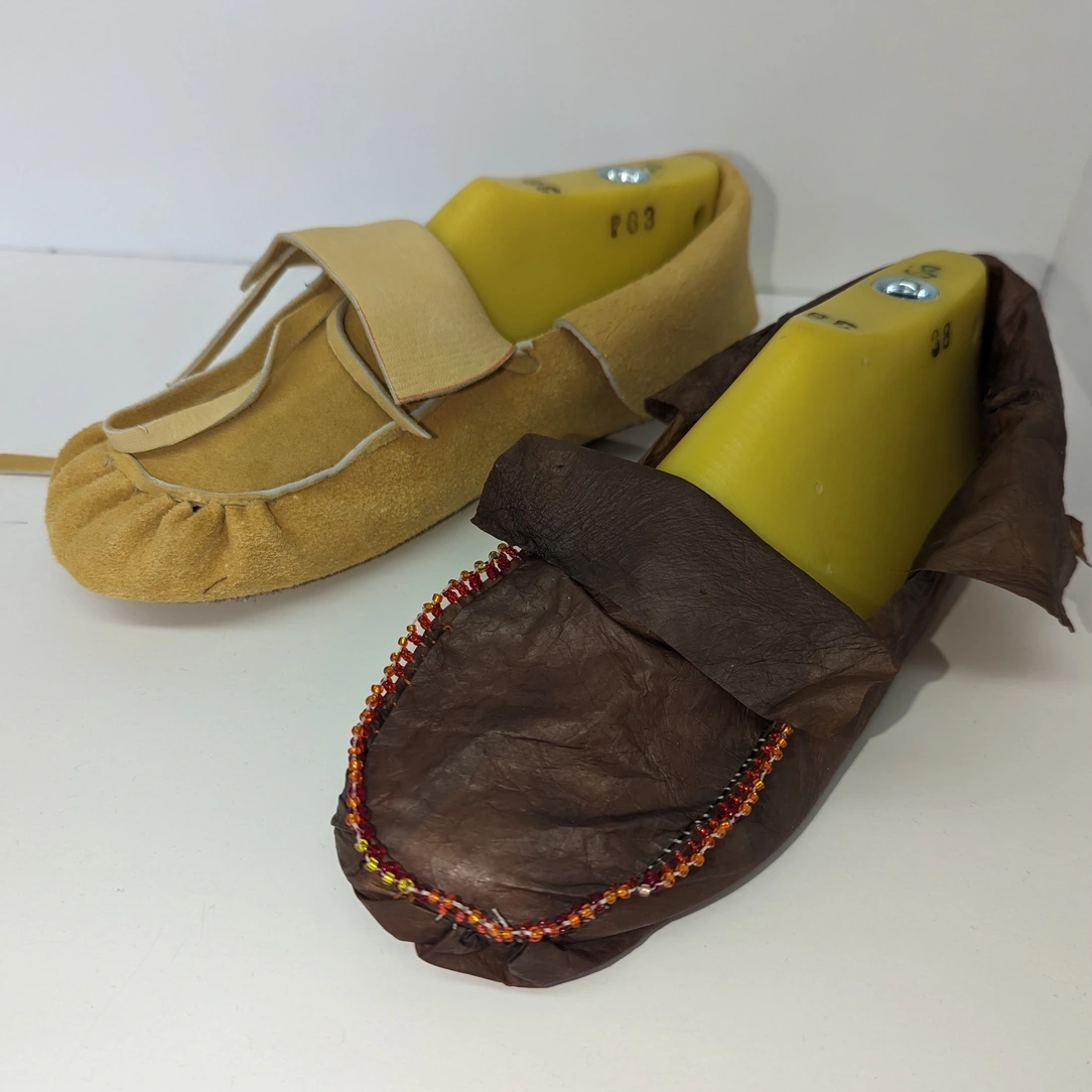 A dark brown SCOBY moccasin with beading sits on a table next to a light brown leather moccasin