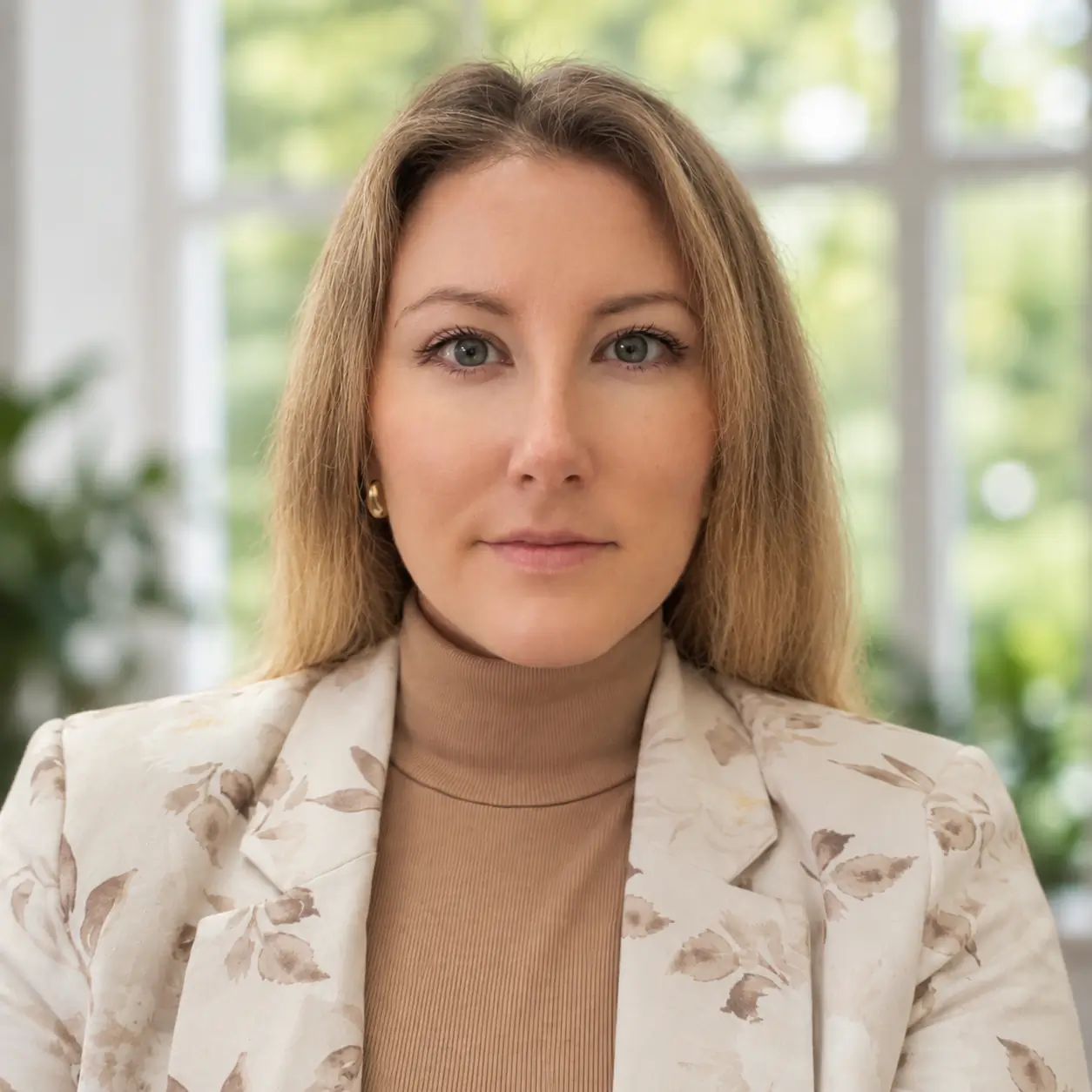 A person wearing a light floral jacket and beige turtleneck, posing in front of a window with greenery outside.