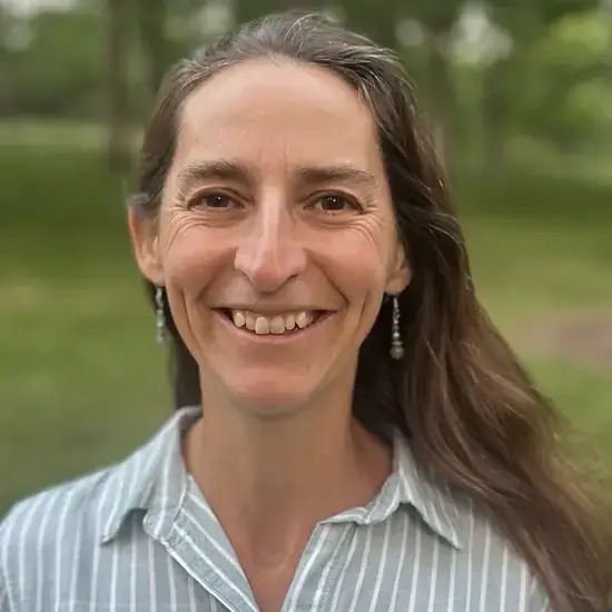 A person smiling outdoors, dressed in a striped shirt and wearing drop earrings, with a blurred green background.