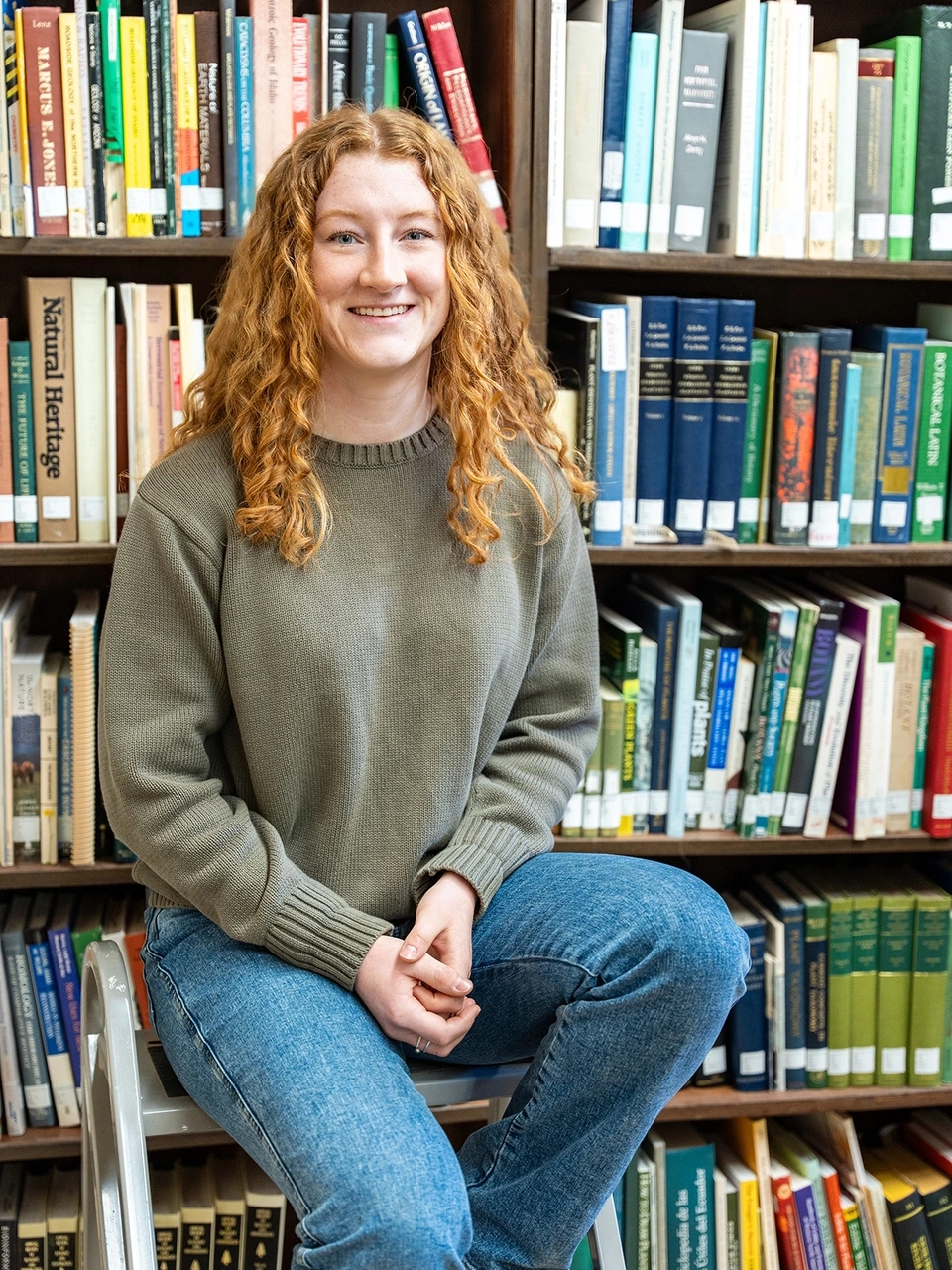 Smiling woman sits on a library stool with rows of books behind her.