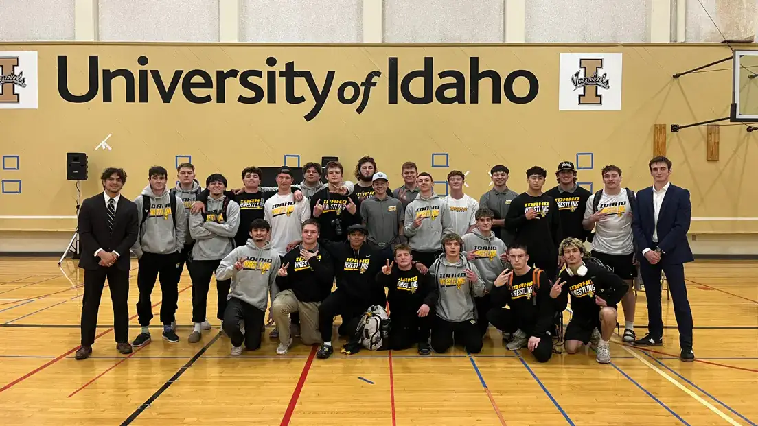 26 men standing or kneeling on gym floor in front of black on yellow University of Idaho sign. 