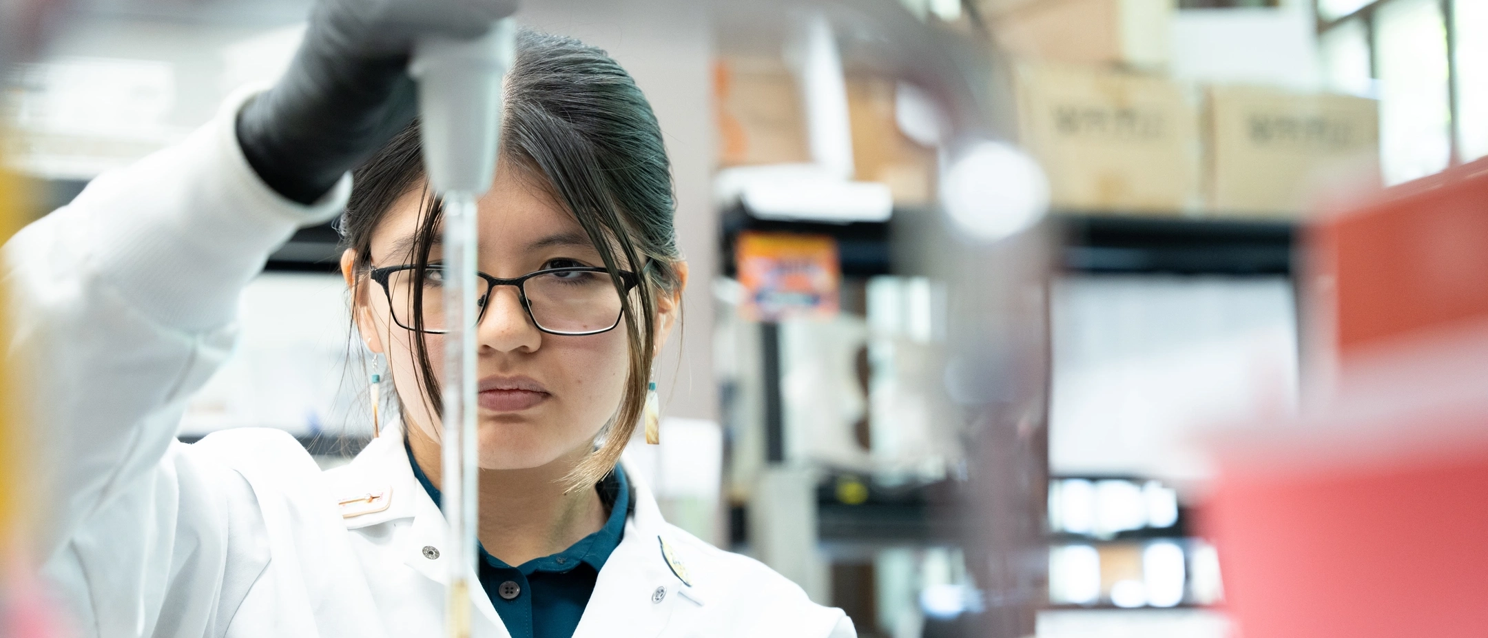 Woman standing behind a shelf full of colorful bottles in lab measures liquid with a pipette