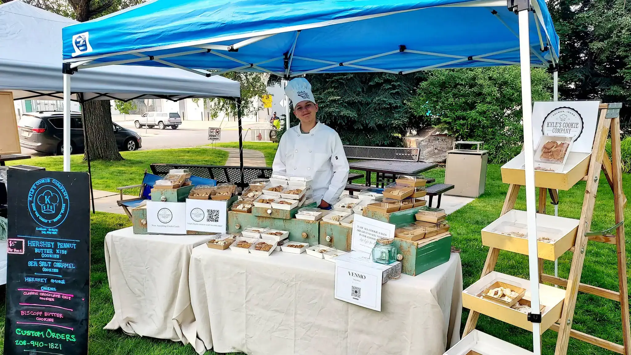 A boy wearing a chef’s hat and jacket poses with hundreds of cookies on table.