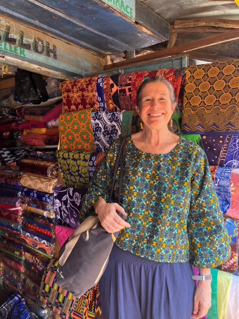 A woman standing in front of colorful printed fabrics in a market setting, holding a bag and wearing a patterned blouse.