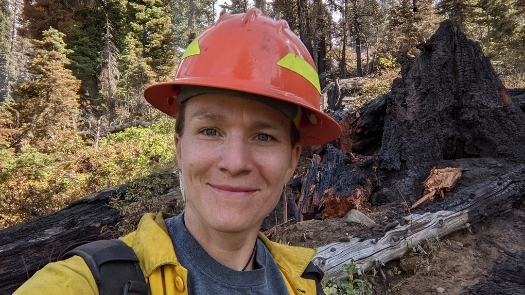 Heather Heward, clad in forest fire fighter gear, smiles in a forest.