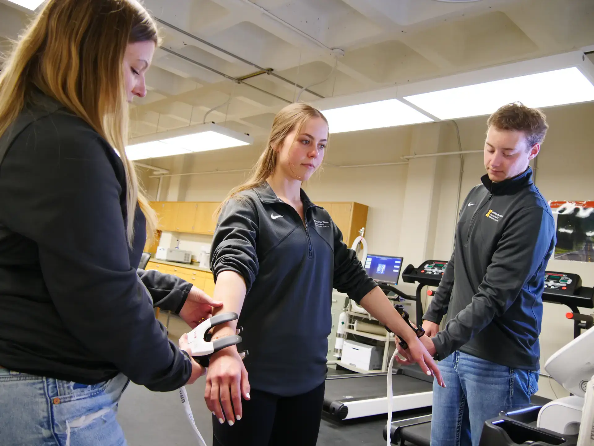 Three individuals in a lab setting using equipment for a physical analysis; one person's arm is being measured.