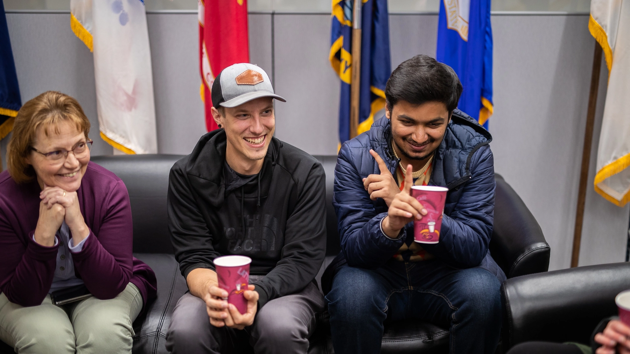 Three people sit on a couch conversing, with international flags behind them.