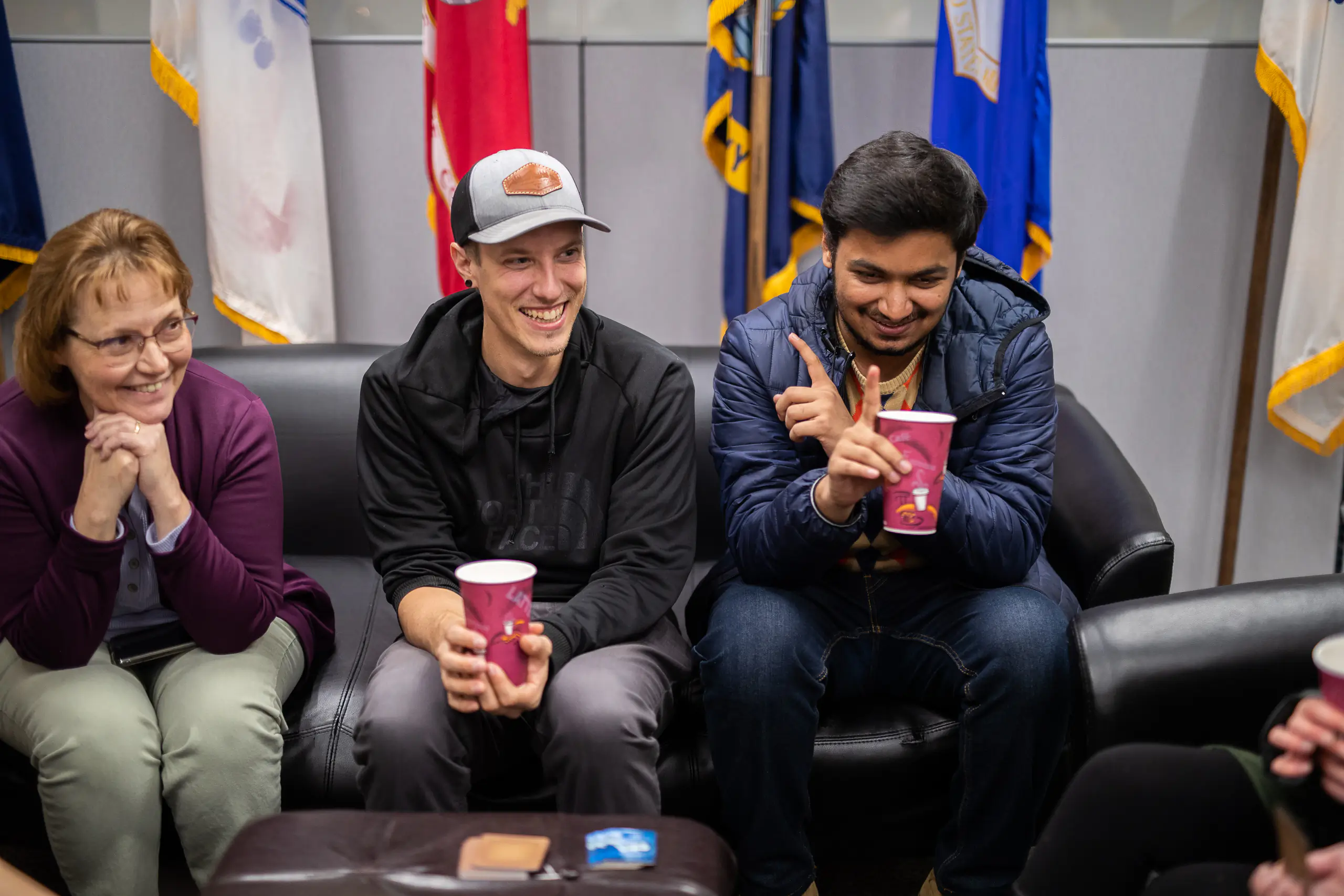 Three people sit on a couch conversing, with international flags behind them.