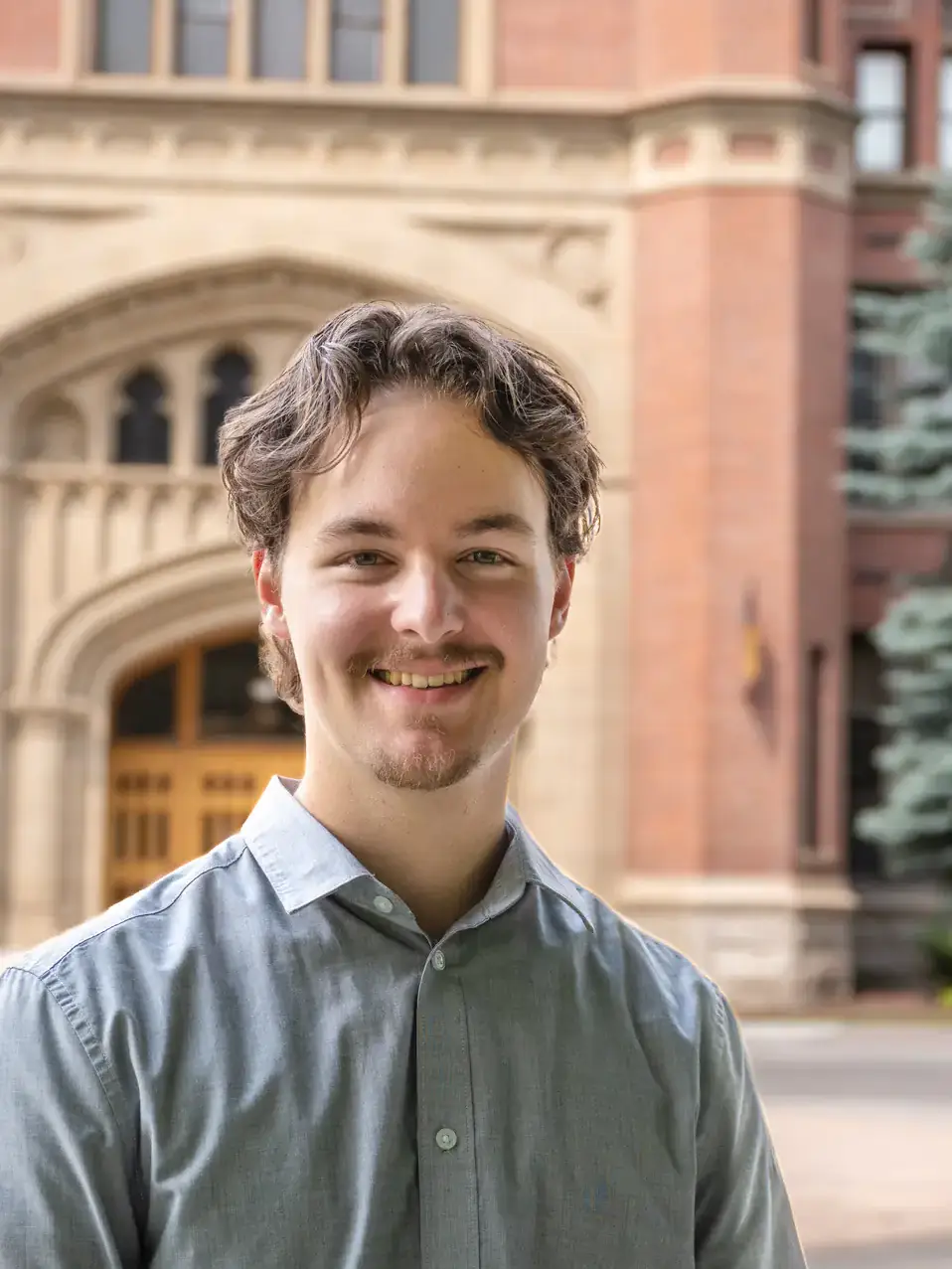 A young individual smiling outdoors, standing in front of a historic brick building with arched windows and a decorated entrance.